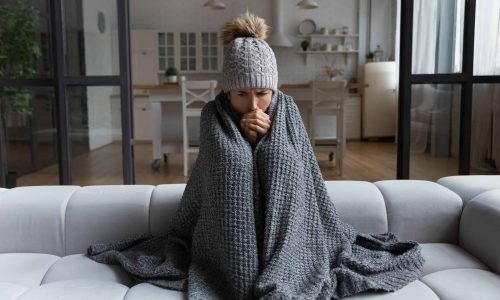 Frozen. Sad latina female sit on couch at freezing cooled studio flat in warm cap and blanket shiver tremble with cold. Unhappy young lady spend time at home feel bad suffer of heating system problems